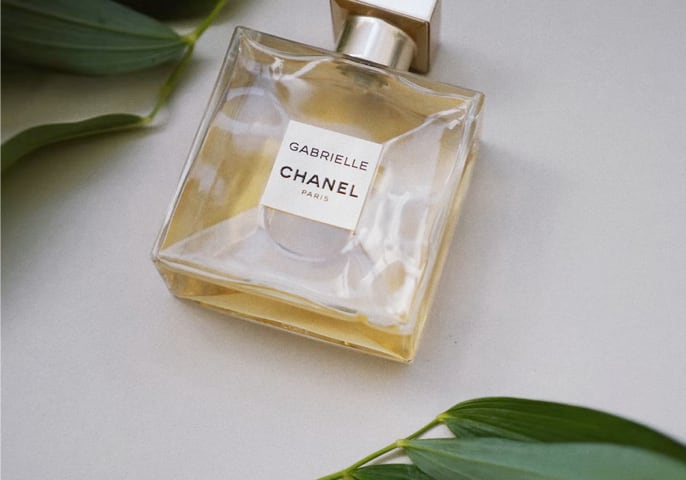 A bottle of Gabrielle perfume, on it's side, ona flat surface, with leaves in the top left and bottom right of the frame